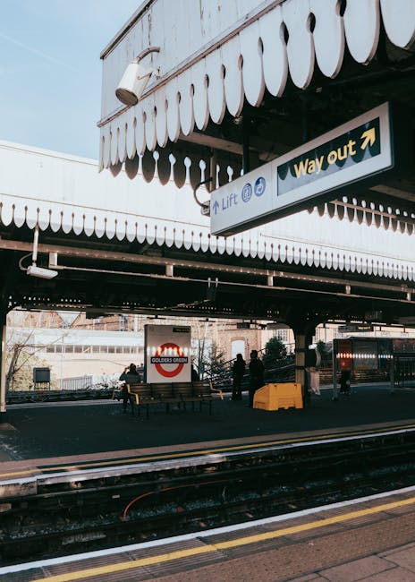 Inside the underground station platform at Maida Vale with tiled walls and flooring, a train approaching in the distance, posters and signs including an illuminated directional sign for the way out, a